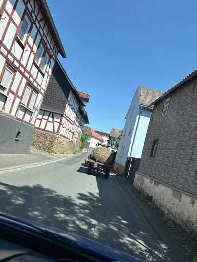A paved road runs through a European village under a clear blue sky. On the left, a traditional half-timbered building with white stucco and red-brown beams is visible. The building has multiple windows with white shutters. A stone foundation supports the lower portion of the house. On the right, a light blue building with several windows stands next to a grey brick building with a textured facade and a weathered section of plaster at its base. Further down the road, another half-timbered building with a red-tiled roof is partially visible. In the middle of the road, a rustic, old trailer with a large, round, metallic tank is being towed. The trailer has two wheels on each side and appears to be moving away from the viewer. Shadows of trees are cast across the road and the side of the blue building. The perspective suggests the image was taken from inside a vehicle. The scene is characteristic of a rural or historic town in Germany, specifically Volpertshausen, given the architectural style.