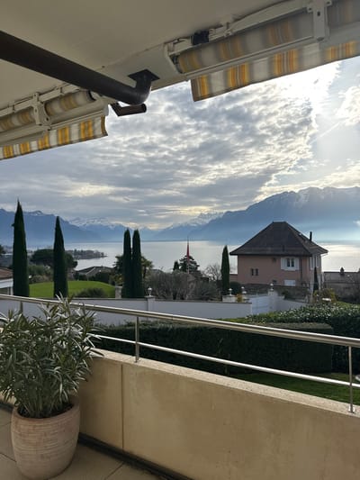 The image displays a picturesque landscape view, captured from what appears to be a balcony or terrace. The image is oriented vertically, but the scene itself is a sweeping horizontal panorama.
In the foreground, on the left side of the vertically oriented image (which would be the bottom if correctly rotated to landscape), are elements of the structure from which the photo was taken. There are two striped awnings, possibly yellow and white or cream, partially extended. Below these, a segment of a light-colored building wall is visible, with a dark, possibly brown or black, downspout running vertically along it and then curving horizontally. On the right side of the vertically oriented image (the right side of a correctly oriented landscape view), a light-colored concrete balcony railing with a silver metal handrail extends across the frame. A large, ribbed terracotta-colored pot holding a bushy plant with silvery-green, narrow leaves is situated on the balcony floor near the railing.
The midground features a well-maintained garden area directly below the balcony, characterized by vibrant green lawns and neatly trimmed dark green hedges. Several tall, slender, dark green cypress-like trees punctuate the landscape. A light pink or beige house with a dark, gabled roof stands out amongst the greenery. Beyond these immediate structures and gardens, a vast body of water, likely a large lake, stretches across the scene, reflecting the sky and light. On the far side of the water, a small red flag, possibly a Swiss flag given the likely location, is visible on a flagpole.
The background is dominated by a majestic range of mountains, many of which are snow-capped on their peaks and upper slopes. The sky above these mountains and the lake is filled with dramatic, bright white and gray clouds, through which sunlight is breaking, creating bright patches and reflections on the water's surface. The overall lighting suggests it is daytime, possibly late morning or early afternoon, with a clear atmosphere providing excellent visibility of the distant features. The scene conveys a sense of serene natural beauty and a well-kept environment. No discernible text is visible in the image.