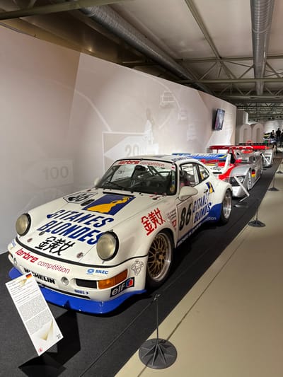 A vintage race car, predominantly white with blue accents, stands on display. Markings include "GITANES BLONDES", the number "86", "larbre compétition", "MICHELIN", and "elf". The front bears a "SILEC" sticker, a German flag, and the words "BAGUES". "100" and "20" are faintly visible on the display surface behind the car. Other vintage race cars are visible in the background. The car is supported by a grey surface. A descriptive card stands near the front of the car.