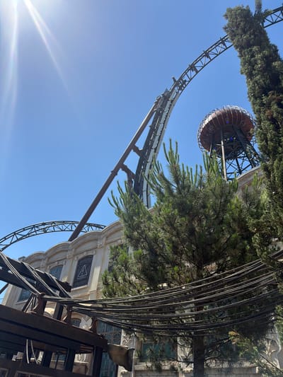 The image shows a roller coaster track curving upwards against a clear blue sky. In the background, a spherical structure with metal accents sits atop a tall, dark tower. To the right, a tall evergreen tree and other foliage are visible. In the foreground, a building with ornate architectural details and arched windows is partially obscured by trees and a structure with numerous dark cables. The roller coaster and the building appear to be part of an amusement park. The building's facade has decorative elements and windows with intricate designs. The overall scene depicts an outdoor amusement park setting on a sunny day, with the roller coaster track being a prominent feature. The presence of the roller coaster and the architectural style of the building suggest it is located in an amusement park. The image does not provide direct information about a specific location in the city of Rust, Germany, beyond the context of an amusement park which is a common attraction there.