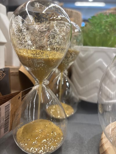 The image shows a close-up of three hourglasses with golden glitter inside, arranged on a shelf. The hourglass in the foreground is partially filled with the glitter, which is falling from the top bulb to the bottom. Another hourglass is visible behind it, and a third hourglass is to the right, only partially in frame. In the background, there is a potted plant with green foliage and a white pot with a zig-zag pattern. The scene appears to be a retail environment, possibly a gift shop or home decor store. The location information provided is irrelevant as there are no visual cues in the image to confirm it is located in Erlenbach am Main, Germany.