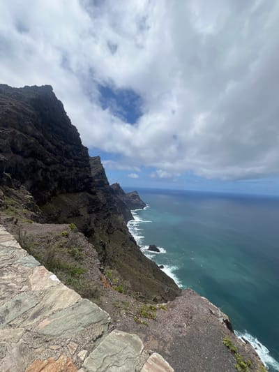 The image shows a view from a cliff in La Aldea de San Nicolás, Spain. The cliff face is steep and rocky, with sparse vegetation. Below, the ocean waves crash against the rocks. The sky is partly cloudy. The perspective is looking down from the edge of the cliff. There are no people visible in the image.