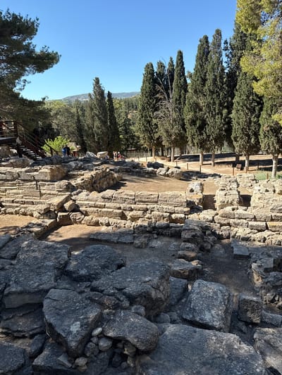 The image depicts the archaeological site of Knossos, located near Heraklion, Crete, Greece. The scene features ancient ruins made of large, rough-hewn stones, forming walls and pathways. In the foreground, weathered gray rocks are scattered across the ground. In the background, several tall, slender cypress trees stand against a clear blue sky, with a hint of hills visible in the distance. A wooden staircase and viewing platform are situated on the left side of the image. A few individuals are visible in the middle ground, exploring the site, with one group clustered near the stairs and another further in the distance. The ground surface is a mixture of dirt and gravel.
