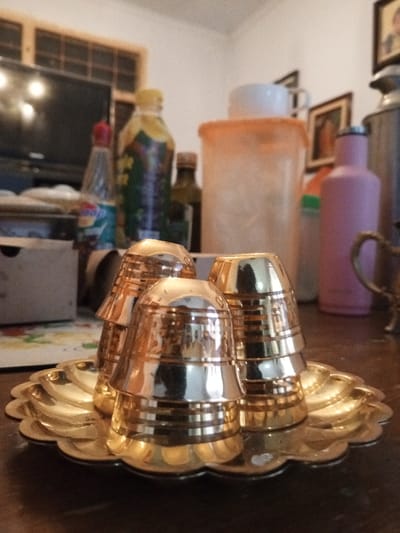 The image displays an indoor scene in Longmiang, Laos. In the foreground, three small, polished golden-colored cups, featuring horizontal ridges, are arranged on a scalloped golden-colored tray. Two of the cups are inverted and stacked, while the third is partially visible. These items rest on a dark, reflective surface, likely a table.

The background is out of focus, revealing several household items. From left to right, a clear plastic bottle with a red cap and a multi-colored label, followed by a taller, opaque bottle with a yellow cap, are present. A section of a dark television screen and a window with a grid pattern are visible to the left. Centrally, a large, translucent orange storage container with a white lid is positioned, topped by a white ceramic mug. To the right, an opaque pink bottle stands next to a smaller, light green bottle. A portion of a decorative metallic object with a handle is visible on the far right. The background wall is light-colored, displaying at least two framed pictures. No individuals are depicted in the scene.