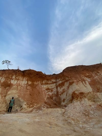 The image displays an active excavation site in Asaga Amangwu, Nigeria, dominated by a large, exposed earth cliff. The cliff face exhibits distinct horizontal stratification, featuring prominent layers of reddish-brown soil interspersed with lighter, whitish to pale yellow soil. The ground in the foreground consists of loose, light-colored earth with several small piles of excavated material near the cliff's base. The sky above is partly cloudy, with a clearer blue area on the left transitioning to a more overcast, lighter sky on the right.

One individual, identified as male, is positioned in the lower-left, standing with his back to the camera at the base of the cliff. He wears a light green or teal short-sleeved shirt with the number "55" visible on the back, and dark trousers or shorts. His left arm is bent at the elbow, while his right arm extends downwards. A second, smaller figure is observed high on the upper edge of the cliff, near the center, appearing to be seated or crouching. A single, small tree with green foliage is visible on the top-left edge of the cliff. A slender, dark object, possibly a digging tool, is partially visible protruding from a small mound of earth to the right of the standing individual.