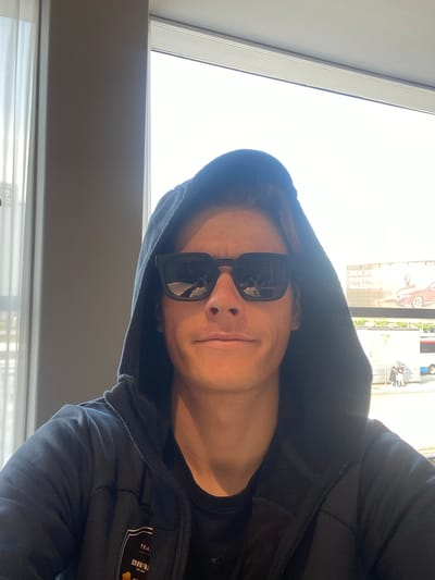 A selfie-style, rotated image shows a person wearing a dark hoodie and dark sunglasses. A window is visible in the background, showing a bright outdoor scene. Reflected in the sunglasses are images of buildings and trees, suggesting an urban setting. A billboard partially visible outside the window features an image of a car and text in what appears to be Danish. The person is indoors, next to the window. The logo on the hoodie reads “DRAI MILANO” and “TEA”. The location is likely in Kastrup, Denmark, based on the billboard signage.
