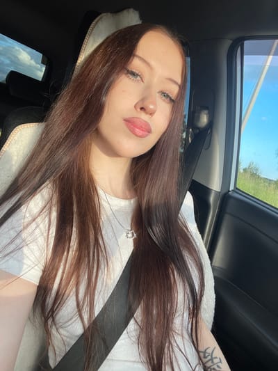 A young woman with long, dark brown hair is taking a selfie while sitting in the passenger seat of a car. The sun is shining brightly, casting shadows and highlighting her features. She is wearing a white t-shirt and a necklace with a small pendant. A seatbelt is visible across her chest. The interior of the car is dark, contrasting with the bright light coming through the car's sunroof, which shows a clear blue sky and green foliage outside. A tattoo is visible on her right arm. The overall mood appears relaxed and casual. The location is identified as Gavrilovka, Russia, and the time of day is likely midday due to the strong sunlight.