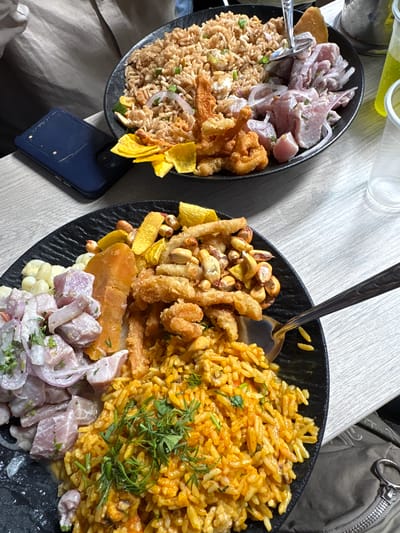 Two large, black oval plates brimming with Peruvian cuisine are presented on a light-colored wooden table, suggesting an indoor dining setting in Jesús María, Peru.

The left plate features a generous portion of chaufa (Peruvian fried rice with visible green onions and red onion rings), accompanied by a large serving of ceviche (diced white fish marinated with red onion), fried calamari or similar seafood pieces, and yellow plantain chips (chifles). A metal serving utensil is visible in the ceviche.

The right plate showcases a vibrant yellow rice dish, possibly Arroz con Mariscos or another type of chaufa, garnished with fresh green herbs. It is surrounded by a variety of fried items, including crispy seafood, toasted corn kernels (cancha or maiz chulpe), and more plantain chips. A large, orange slice of sweet potato (camote) and white corn kernels (choclo) are visible next to a portion of ceviche topped with red onion. A metal spoon rests in the yellow rice.

In the upper left background, several clear glasses or bottles containing a light-colored liquid are present. A dark blue smartphone with a textured case is partially visible on the table near the bottom left, next to the left plate. A grey bag or jacket with a metallic zipper pull can be seen in the upper right corner. The bright and even lighting suggests it is daytime. No discernible text is visible in the image.