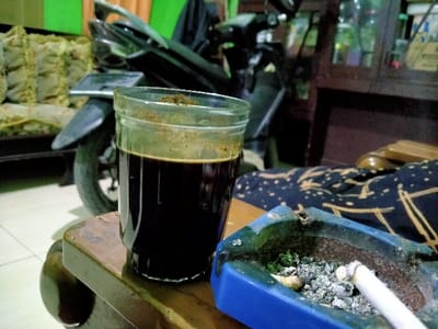 A glass of black coffee sits on a wooden table next to a blue ashtray. The ashtray contains cigarette butts and ash, with a lit cigarette resting on the edge. A parked black motorcycle is visible in the background, along with cushioned seating and a cabinet. The lighting is dim, casting shadows across the scene.
