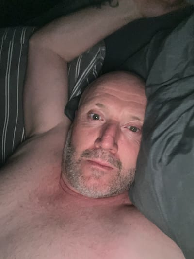 A man lies in bed under a gray comforter, his head visible above the covers. He has a shaved head, stubble, and light-colored eyes. The bed has striped bedding. The scene is indoors with low lighting.
There is no information to suggest that the location is in the city of Sint-Niklaas, Belgium.