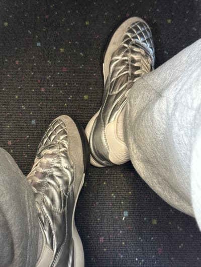 An overhead photograph captures the lower extremities of an individual, likely seated, within an interior environment. The subject's feet are clad in silver athletic shoes featuring quilted metallic uppers, light gray suede overlays, white midsoles, and black outsoles. Light gray sweatpants are worn, partially obscuring the ankles. The footwear rests on a dark gray carpet patterned with small, uniformly distributed multi-colored squares in pink, blue, green, and yellow. Visual evidence in the image does not permit determination of a specific location within Tijuana, Mexico.