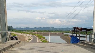 A wide-angle, eye-level shot shows a view from a concrete bridge in Baroh Lancok, Indonesia. The bridge has a light-colored concrete surface with a curb. To the left, a metallic pole and railing are visible, with a dark-colored motorcycle parked on the bridge.

The scene opens to a landscape with a winding paved road on the left, bordered by a rocky embankment and green vegetation. Beyond the road, a body of calm water, possibly a river or inlet, stretches into the distance. Several small boats are visible on the water. On the right side of the water, a small, open-air shelter with a rusty corrugated metal roof and wooden posts stands, partially covered by a blue tarp. The background features a dense green treeline and rolling hills, with larger mountains in the far distance under a cloudy sky. Power lines are strung across the upper portion of the image. The overall impression is a rural, coastal environment under an overcast sky.