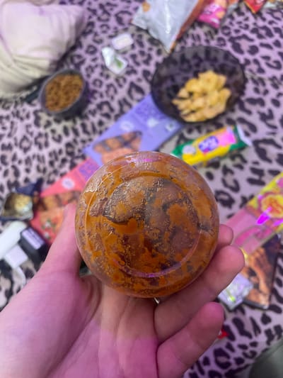 A hand is holding a clear plastic container filled with a reddish-brown, chunky substance. The background is a leopard-print fabric, on which several snack food wrappers and containers are scattered. These include a dark bowl with yellow chips, a small container with what appears to be a white dip and green herbs, and various colorful packages. The scene is dimly lit, with pink and purple hues suggesting artificial lighting. The location is described as the city of Zmeinyi, Russia.