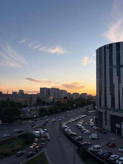 This is a high-angle, vertical shot of a city scene at dusk. The sky is a gradient of light blue to soft orange, with wispy clouds. In the foreground, a modern, curved building with a striped facade dominates the left side of the frame. Below the building, a multi-lane road curves through the cityscape, with numerous cars of various colors, including white, black, and red, driving and parked. Several buildings, some with multiple windows and balconies, are visible in the midground and background, interspersed with trees. The lighting suggests the sun is setting or has just set, casting a warm glow on the clouds and the side of the road. The overall atmosphere is urban and somewhat busy, with the flow of traffic being a prominent element. No people are clearly visible, but the presence of cars indicates ongoing activity. Some signage is visible on the buildings, but it is too small to be legible. The scene appears to be a typical city street and parking area in a residential or commercial district.