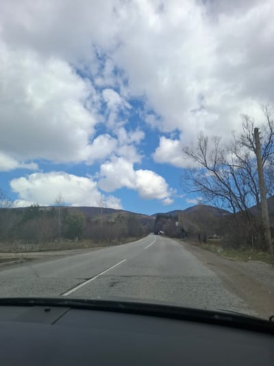 A view from inside a vehicle looking towards a paved road in Gintsi, Bulgaria. The road has a white dividing line. To the left of the road, there are barren trees and a cloudy, blue sky. To the right, there is a wooded hillside. The vehicle's interior trim is visible on the right side of the frame.