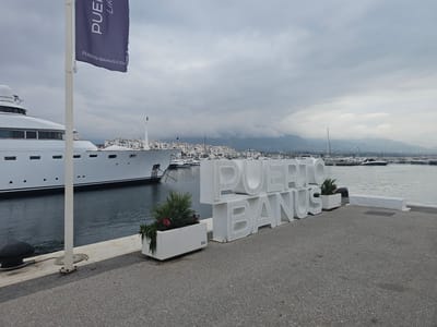 The scene shows a gray concrete pier in Puerto Banús, Nueva Andalucía, Spain. A large, white yacht is docked on the left side of the pier, with a flag pole and a navy-colored flag bearing the text "PUERTO-BANUS.COM" in white. A large, white, three-dimensional sign spells out "PUERTO BANUS." Rectangular white planters filled with green plants frame the sign. In the background are white buildings, more yachts, and a gray, cloudy sky.