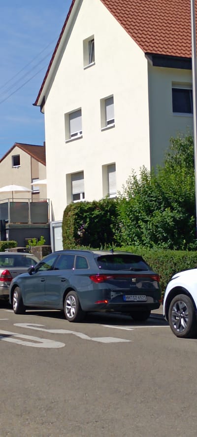 In Schwaikheim, Germany, on a sunny day, a dark grey station wagon with license plate "HH SZ 6521" is parked on a street. The car is facing right and is positioned in the foreground, partially obscuring a white car on its right. The station wagon has its rear taillights illuminated. Behind the vehicles, a dense green hedge runs horizontally across the scene. Further back, a light-colored building with a red-tiled roof and several windows is visible, with one window open. A smaller, adjacent building with a light beige facade and a red roof is also present. The sky is clear and blue.