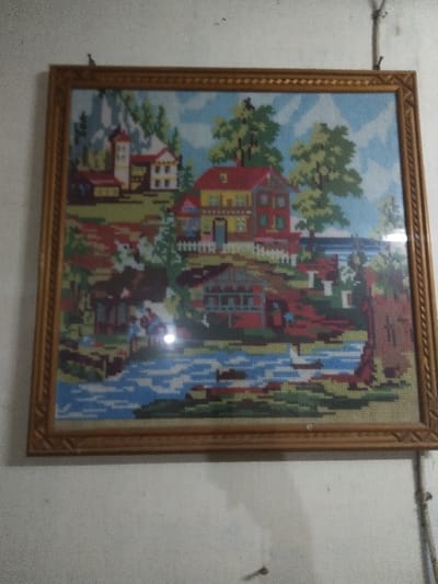 A framed, rectangular needlepoint or cross-stitch artwork hangs centered on a plain, light-colored wall. The frame is light brown or gold-colored wood with an ornate, carved pattern along its inner edge. The artwork depicts a colorful, idyllic landscape scene in a blocky, pixelated style, visible through a glass cover that exhibits a central glare point.

The scene features a river with blue water and white ripples flowing horizontally across the lower third of the image. On the left bank, two small figures, one appearing to be bending, are situated near the water. Behind them, a small brown-roofed building is nestled among green and brown terrain. A prominent, multi-story house with a red roof, yellow walls, and brown trim occupies the central mid-ground, fronted by a white picket fence. Various green trees surround this house. To the left, a larger, castle-like structure with a red conical tower roof and light-colored walls stands on a green hillside with visible rock formations. Additional green trees are scattered across the landscape, which is set against a light blue sky with white cloud-like elements. On the right bank of the river, a smaller red-roofed building is partially visible, flanked by trees.

The wall surface is off-white or light grey, showing minor imperfections and a slightly textured finish. Two small hooks or nails are visible along the top edge of the frame, securing it to the wall. A dark, thin vertical line, possibly a cable or wire, is affixed to the wall on the right side of the frame. A single dark screw head or nail is present in the lower central portion of the wall, below the frame. The image does not contain identifiable visual information to specify its location within the city of Chimay, Belgium.