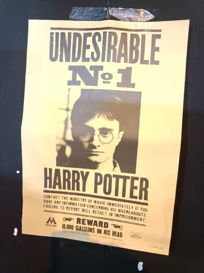 A light-yellowish, vertical poster is affixed to a dark, matte background surface in El Port De Sagunt, Spain. The poster displays black text: "UNDESIRABLE Nº1" at the top, followed by a black and white portrait of a young male with dark hair and round glasses. Below the portrait, the text "HARRY POTTER" is prominently featured. Further text states: "CONTACT THE MINISTRY OF MAGIC IMMEDIATELY IF YOU HAVE ANY INFORMATION CONCERNING HIS WHEREABOUTS. FAILING TO REPORT WILL RESULT IN IMPRISONMENT." At the bottom, a reward offer is visible: "REWARD 10.000 GALLEONS ON HIS HEAD", accompanied by pointing hand graphics and a stylized "M" logo. The poster is secured at its top edge by two pieces of dark tape, with a small reflective element present near the right piece of tape. Several small white specks are distributed on the dark background surface, notably on the left side.