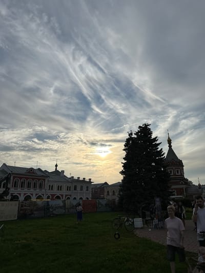 This is a medium-shot, eye-level, outdoor photograph taken in the city of Putyatino (Semivragovskaya S/A), Russia. The scene captures a broad view of a public space with several buildings, a large evergreen tree, and a portion of the sky.
In the background, a long, multi-story building with red and white facades is visible. The architecture features arched windows and decorative elements. To the right of this building, a portion of a church or religious building with a red brick lower level and a domed, golden-topped roof is present. The sky above is dramatic, filled with wispy, elongated clouds that stretch across the frame. The sun is setting or rising, creating a bright, hazy glow towards the upper right quadrant of the image.
In the mid-ground, a tall, dark green evergreen tree stands prominently, silhouetted against the sky and the buildings. In front of the buildings, there is a grassy area. A fence or barrier with large printed banners depicting various scenes, possibly historical or artistic, runs along the edge of the grassy area.
In the foreground and mid-ground, there are several people. On the far left, a person is standing on the grass facing away from the camera, wearing a light-colored top and a blue skirt. Towards the right, a group of people are walking on a paved path or area. One woman in the foreground is wearing a pink t-shirt and dark shorts, walking towards the camera. Another man in a white t-shirt is visible to her right, also walking. Other individuals are present in the background, some appearing to be interacting or browsing displays. A bicycle is parked on the grass to the left of the paved area. Several stacks of what appear to be white crates are also visible near the bicycle.
The overall atmosphere is that of a public gathering or event, possibly a park or historical site, during a time of transition in the day, indicated by the sky and sunlight.