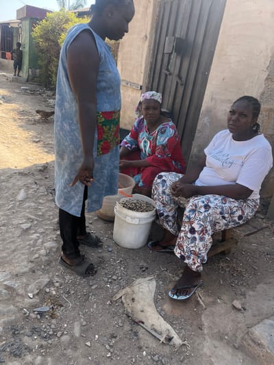 The image depicts an outdoor scene in Dzhidu, Nigeria, featuring three adult females and a partial view of a fourth individual.
In the foreground, a dark-skinned female stands on the left, oriented rightward, wearing a light blue-grey sleeveless tunic with a floral pattern panel at the waist, black trousers, and dark slide sandals. Her right hand is lowered, with the index finger extended downwards. She appears to be interacting with two dark-skinned females seated on the ground to her right.
The central seated female wears a multi-colored patterned garment in red, blue, and white, along with a light-colored floral headscarf. She is positioned on a low surface, looking towards the standing female. To her right, the third female is seated on a low wooden stool, wearing a white t-shirt with the text "I AM FREUND (ICH)" and white trousers patterned with blue, red, and brown motifs, along with black flip-flops. Her hands are clasped in her lap, and her gaze is also directed towards the standing female.
Between the standing and seated individuals, two white plastic buckets filled with small, light brown, uniform-sized items, possibly nuts or seeds, are visible on the unpaved, gravelly ground. A woven basket is partially visible behind the buckets. In the immediate foreground, a torn, light-colored piece of material, possibly plastic or fabric, rests on the ground.
The background features a light-colored wall with a dark wooden door or gate behind the seated women. Further back on the left, a green structure and some foliage are visible, along with a partial view of another individual, possibly a child, standing near the green structure. A small fowl, likely a chicken, walks on the dirt path to the left of the standing female. The scene is illuminated by direct sunlight, casting visible shadows.