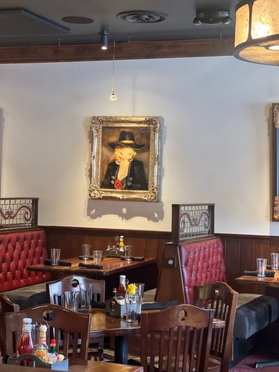 The image shows the interior of a restaurant in Jackson, United States of America. The restaurant has a warm and inviting atmosphere with dark wood paneling, red leather booths, and wooden chairs. A painting of a woman in a black hat and jacket with a red rose is displayed on the wall, adding a touch of elegance to the space. The tables are set with glasses, napkins, and condiments, indicating that the restaurant is open for business. The lighting is dim and ambient, creating a cozy and intimate dining experience.