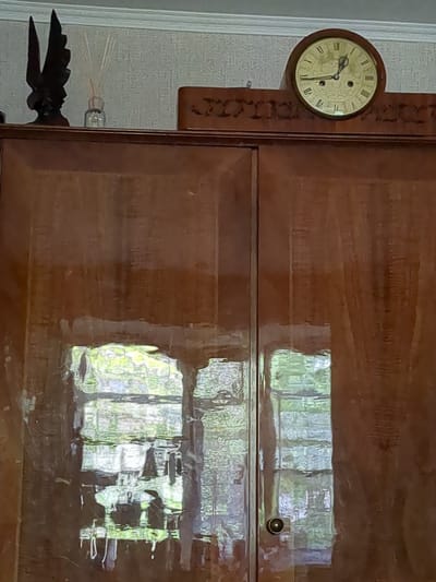 A wooden cabinet with a polished, reflective surface stands against a lightly textured wall. On top of the cabinet, a dark, wing-shaped sculpture resembling a bird is positioned to the left. Next to it, a small glass vase holds several thin, light-colored reeds, likely for aromatherapy. Further to the right, an ornate, dark wooden decorative piece with carved patterns is visible. Centered on the top of the cabinet is a round, vintage clock with Roman numerals. The clock face is light yellow, and the hands indicate a time around 10:10. The cabinet itself has two doors, each with a subtle sheen that reflects the light and the surrounding environment, including what appears to be a window with panes and greenery outside. A round, brass-colored doorknob is present on the right-hand door. The image appears to be taken indoors, likely in a residential setting, with no indication of the specific location in Boryspil, Ukraine.
