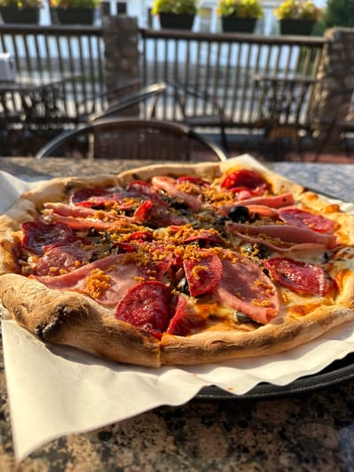 A close-up, eye-level shot captures a gourmet pizza topped with pepperoni, ham, olives, and crispy fried onions. The pizza rests on a white paper liner atop a black pizza tray, which is placed on a speckled brown granite table. In the blurred background, an outdoor patio setting is visible, with wicker chairs, a railing, and planters containing green foliage. The lighting suggests a sunny day, casting shadows and highlighting the textures of the pizza. This scene is taking place at a restaurant in Rūdiškės, Lithuania.