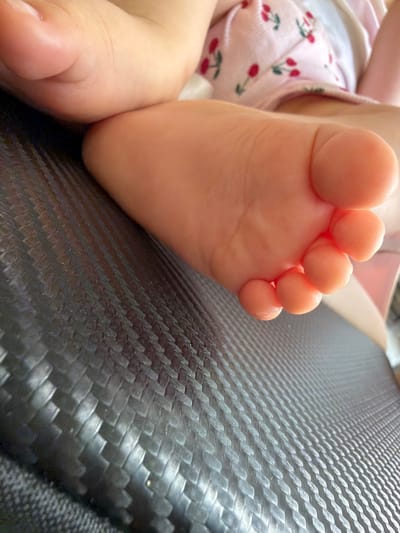 The image is a close-up shot of a baby's foot, with the toes and sole clearly visible. The foot appears to be resting on a textured, dark surface, possibly a chair or sofa. The baby is wearing pink shorts or pants with a cherry pattern. The background is slightly blurred, suggesting a shallow depth of field, and includes a hint of another body part, likely belonging to the same baby. The lighting highlights the soft texture and color of the baby's skin. There is no indication or presence of the city of Rusokastro, Bulgaria in this image.