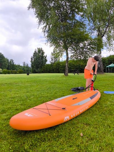In Vecumnieki, Latvia, on a grassy field under a cloudy sky, a young person wearing orange shorts is actively inflating an orange stand-up paddleboard with a red hand pump. The paddleboard, labeled "Aqua Marina," lies on the grass in the foreground. Beside it is a black backpack. In the background, trees and a green gazebo are visible, suggesting an outdoor recreational area.