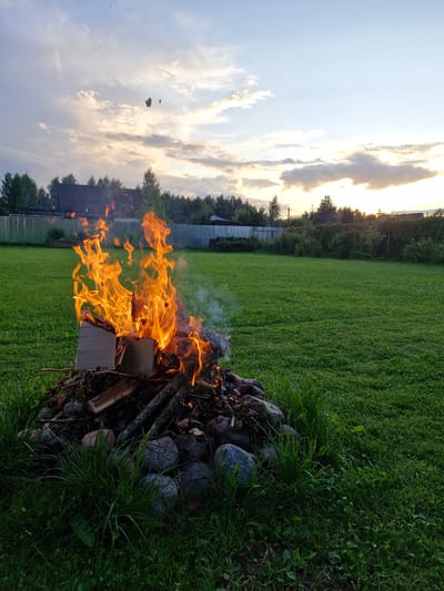 The image depicts an outdoor scene at dusk, likely in the village of Sorochkino, Russia. A bonfire burns vigorously in the foreground, composed of branches, logs, and some cardboard material. The flames are bright orange and yellow, with a plume of white smoke rising from the fire. The bonfire is surrounded by large, round stones and grass. Behind the bonfire, a well-maintained green lawn stretches out, bordered by a metal fence and a dense line of trees. In the distance, a residential building with a dark roof is visible, partially obscured by the trees. The sky above is a mix of pale blue and grey clouds, with the warm hues of the setting sun visible on the horizon. Two small, dark objects appear to be falling from the sky in the upper portion of the image, though their nature is unclear. The overall impression is of a peaceful, rural evening setting.