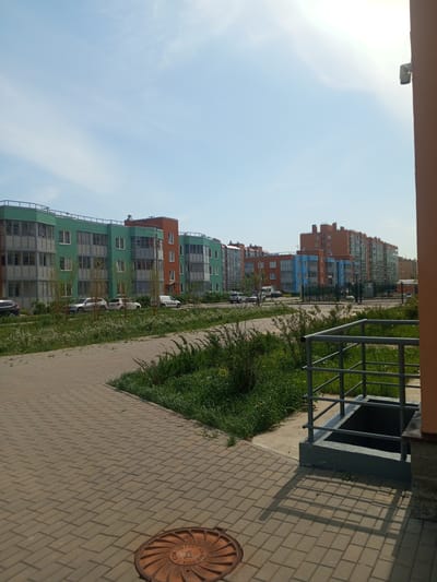 The image depicts a street scene in Kommunar, Russia, showcasing a row of colorful, modern apartment buildings. The buildings are painted in shades of green, orange, and blue, with multiple stories and visible balconies. Several parked cars are visible in front of the buildings.
A paved brick walkway is in the foreground, leading into the scene. To the right, a set of metal railings surrounds a sunken area, likely for utility access. Patches of green grass and foliage are scattered throughout the scene. A manhole cover is visible on the left side of the walkway. The sky is clear and blue, suggesting a sunny day. The overall atmosphere is calm and residential.