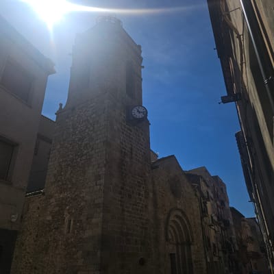 A ground-level view of the city of La Jonquera, Spain, in daylight showcases a building made of tan stone and mortar construction. The building has a prominent tower affixed with a clock. The clock is high up on the building's surface and has a white clockface with Roman numerals and black hands. The tower is square and stacked with blocks of stone. A clear sky can be seen around the tower. The building's architectural style is Gothic, with a rounded arch over the entrance. A street flanks each side of the building, with other buildings parallel to the pictured one. The sun is above the scene, with the building primarily in shadow.