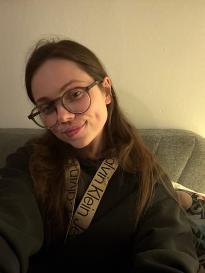 A woman with long brown hair and round glasses is lying on a gray couch, wearing a black Calvin Klein sweatshirt. She is positioned against a light beige wall. The location is not discernible from the image.