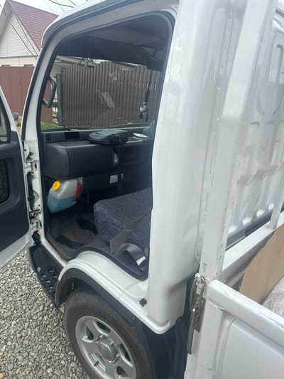 The image shows the interior and exterior of a white utility truck, viewed from a slightly elevated, side angle. The truck appears to be parked outdoors on a gravel surface, with residential buildings and a reddish-brown tiled roof visible in the background, suggesting a semi-rural or suburban setting in Ptichnik, Russia. The sky is overcast, implying a cloudy day.

Inside the truck's cab, two black seats are visible, with the passenger seat partially covered by a patterned blue and grey fabric. A plastic container filled with a light blue liquid and a yellow cap sits on the floor behind the passenger seat. The dashboard and part of the steering wheel are visible. The truck's door is open, revealing the interior.

The exterior shows the side of the truck, including the wheel well with a silver alloy rim and tire. The truck bed is also visible, appearing to be made of white metal. A metal latch mechanism is attached to the side of the truck, likely for securing the cargo area. The overall impression is of a work vehicle in a residential environment. There is no visible text in the image.