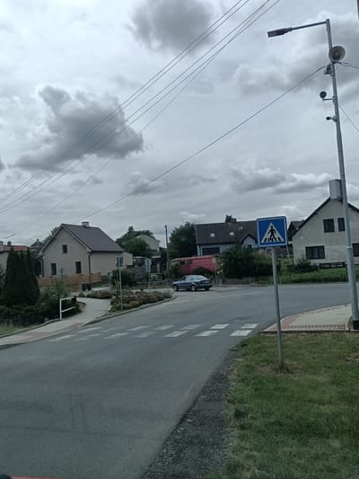 The image depicts a cloudy day scene in the city of Čistá, Czechia, focusing on a street intersection with a pedestrian crossing. A dark blue sedan is seen driving across the pedestrian crossing. To the right of the crossing, a blue and white pedestrian crossing sign is mounted on a pole. Behind the sign, a building with dark windows is visible. A street lamp with multiple attached devices, including what appears to be a speaker and cameras, is also present on the right side of the image. The road surface is asphalt, with white markings for the pedestrian crossing. Beyond the road, there are houses, trees, and a paved walkway. The sky is overcast with dramatic, dark clouds.