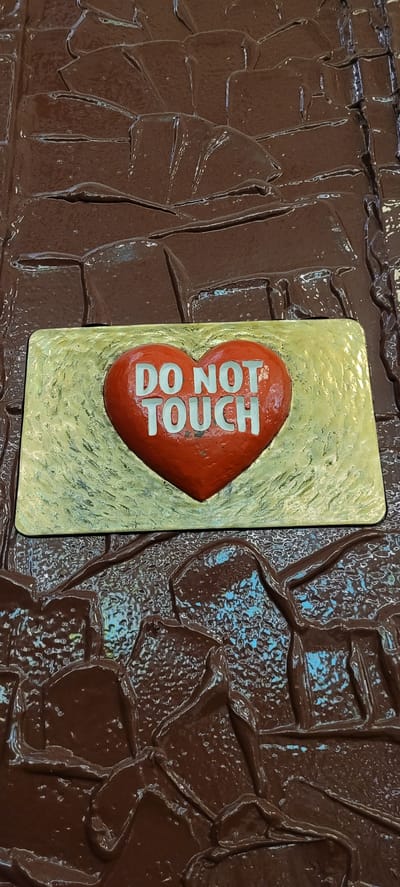 The image shows a close-up of a red heart-shaped plaque with the words "DO NOT TOUCH" in white letters. The plaque is mounted on a textured, golden-colored surface. The background appears to be a dark, glossy, textured material, possibly resembling melted chocolate or a similar confectionery. The overall scene is presented in a vertical orientation, with the plaque positioned in the lower half of the frame.
This object is located in the city of Wattens, Austria, specifically within the Swarovski Crystal Worlds (Swarovski Kristallwelten). The Swarovski Crystal Worlds is a museum and entertainment complex dedicated to the art and heritage of Swarovski crystal. The "DO NOT TOUCH" heart is a decorative element within the artistic installations found at the site.