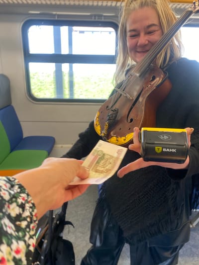 A woman with blonde hair holds a violin in her left hand and a bank card reader in her right hand. Another hand is offering the woman a 100-ruble bill. This image was taken inside a train or bus. Novoivanovskoe, Russia is not visible.