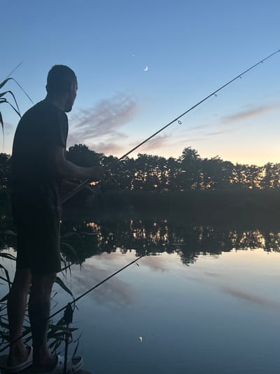 The image depicts a silhouette of a person fishing in a body of water. The person is facing away from the camera, looking towards the water and the opposite shore. They are holding a fishing rod, casting it towards the water. The scene is set by a tranquil lake or river with reflections of trees and the sky on the water's surface. The sky is clear with some wispy clouds, and a crescent moon is visible. The time of day appears to be either early morning or late evening, as indicated by the soft lighting and the colors in the sky. The overall mood is peaceful and serene, suggesting a moment of quiet contemplation and enjoyment of nature. The location is identified as Platnirovskaya, Russia. No text is visible in the image.