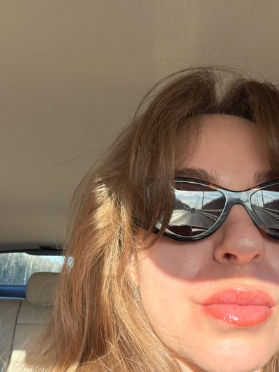 The image shows a close-up, slightly rotated view of a person's face wearing sunglasses. The person has light-colored hair and shiny lips. The sunglasses reflect a cityscape, possibly the exterior environment of Давыдково, Russia. The scene is well-lit, with sunlight highlighting the face and hair. The background is a light-colored, possibly textured surface.