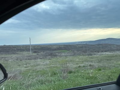 The image depicts a rural landscape observed from a vehicle, with parts of the dark window frame and a side mirror visible in the foreground. The scene is located in the area of Suhache, Bulgaria. The immediate foreground consists of a grassy field, displaying a mix of vibrant green and dried, lighter-colored vegetation. Scattered throughout this field are sparse, bare shrubs and individual blades of long grass.
In the midground, rolling hills are present, predominantly covered with dormant, grayish-brown brush and leafless trees. A series of utility poles, each supporting multiple horizontal power lines, traverses the midground from left to right. At least five such poles are clearly identifiable. The distant background features more undulating terrain, with a prominent hill on the far right exhibiting a structure resembling a communication tower.
The sky is largely overcast with dense, grey cloud cover, though a brighter, lighter blue-white band is visible along the horizon line on the right, suggesting indirect or diffused light. No people, animals, or specific actions are discernible within the frame.