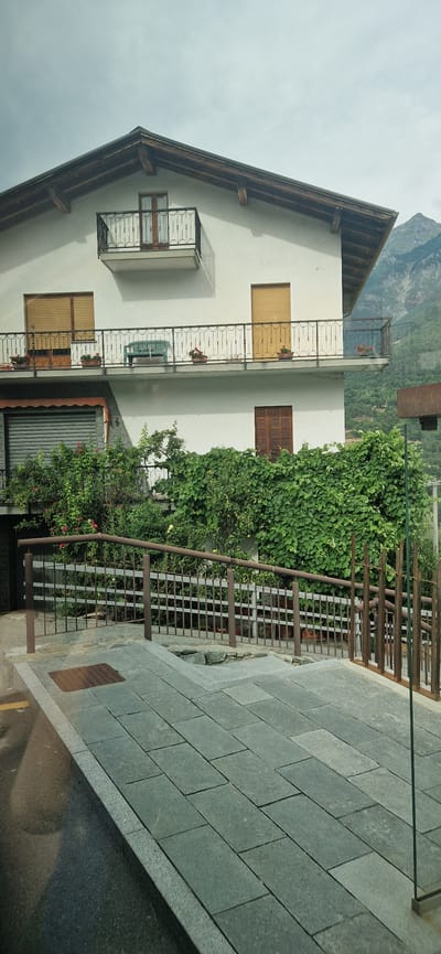 The image shows a white multi-story building with a dark wooden roof and a dark metal balcony railing. The building has multiple windows with wooden shutters and a dark wooden door on the lower level. There is lush green vegetation surrounding the lower part of the building, including climbing vines and flowering plants. A metal fence runs along the foreground, separating the building from a paved area made of large gray stone tiles. In the background, a mountain range with green slopes and a cloudy sky are visible. The location is identified as Challand-Saint-Victor, Italy. There are no people or specific actions depicted in the scene.