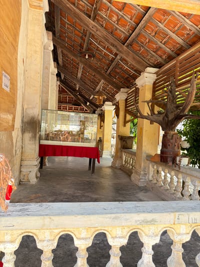 The image shows an outdoor walkway with a covered roof supported by pillars. The roof is made of terracotta tiles and wooden beams, showing signs of age. The walkway has a decorative railing with balusters. On the left, there is a display case with a model inside, covered by a red cloth on a table. Further down the walkway, there is a large, gnarled piece of wood mounted on a wooden base, with hanging planters. The walls are painted yellow and show wear. The ground is paved. A sign with Vietnamese text is attached to the left wall. The scene appears to be in Hội Tín, Vietnam, possibly a historical site or temple. There are no people visible in the image.