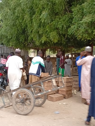 A group of people gather around stacks of cardboard boxes. Some individuals wear traditional attire. A two-wheeled cart stands near the boxes. A motorcycle is visible on the left. The scene takes place outdoors, possibly in a market, with a large tree overhead. The ground appears to be dirt or gravel.