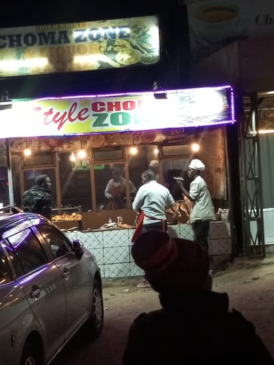 The image displays a night scene in Kinanie ward, Kenya, centered on an illuminated street-side food establishment. In the foreground, the back of an individual wearing a dark garment and a dark hat with a red band is visible, observing the establishment. A silver sedan is parked on the left, reflecting the neon lights.

The establishment features two prominent signs: an upper sign reads "HIGH POINT CHOMA ZONE & LIQUOR," depicting food items, and a lower sign, "Style CHOMA ZONE," outlined with purple LED strips and displaying "24/7 WELCOME." Below the lower sign, the front of the eatery is lit by several bulbs, revealing food counters and personnel.

Four individuals are identifiable near or within the establishment. On the left, a man in a dark, possibly camouflage-patterned jacket stands near the parked car. Inside or directly at the counter, three male workers are active: one partially visible in a light-colored apron, another in a light shirt and red apron attending to items on a counter, and a third in a white chef's hat and light uniform holding a plate or tray. Trays containing what appears to be grilled meat are displayed on the counters. The lower facade of the building is tiled with a white and light-colored diamond pattern. The ground is unpaved dirt or gravel. A metal support structure and a white appliance are visible on the right side of the scene.
