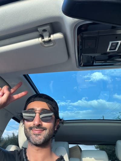 A man is captured in a close-up selfie from inside a vehicle, with the clear blue sky and scattered clouds visible through the sunroof. He is wearing sunglasses and a black baseball cap, and he is making a peace sign with his left hand. The interior of the car is visible, with the car's roof lining and part of the passenger seat. The scene appears to be set in a suburban or rural area, as indicated by the glimpse of trees and power lines in the background. This image could have been taken on a road in South Staffordshire, United Kingdom, given the natural light and visible vegetation.