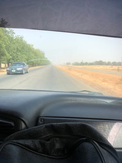The image depicts a road scene in Charanchi, Nigeria, captured from the interior of a vehicle looking forward through the windshield. The lower foreground is occupied by a black, textured dashboard, on which a black bag with a repeating embossed pattern rests. The vehicle's windshield displays a prominent curved white crack or scratch across its upper central area, along with other minor imperfections; the top portion of the windshield is tinted.

The scene outside features a two-lane paved road extending into a hazy distance, flanked by a dense row of green trees on the left side. To the right of the road is a wide, dry, reddish-brown dirt shoulder. A dark-colored sedan, consistent with a Honda model, is visible in the opposing lane on the left, traveling towards the camera. A light-colored object is placed beneath its driver's side windshield wiper, and a green license plate is attached to its front bumper. Further down the road on the right dirt shoulder, a solitary individual, wearing a red top and dark lower garments, stands near a red cylindrical object, facing away from the camera. Utility poles are sparsely distributed along the right side of the road in the far distance. The overall lighting is bright, with significant atmospheric haze reducing visibility.