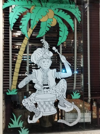 A whimsical window painting features a palm tree with coconuts above a dressed monkey playing a drum. The monkey wears a turban with a feather, a vest, and curled shoes, with a dotted pattern on its pants and tail. Green, stylized foliage accents the lower portion of the design. The background consists of horizontal blinds and reflections of an interior space.