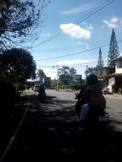 The scene, set in the city of Ka Thaung (Upper), Myanmar, captures an outdoor road junction under a bright, clear blue sky with scattered cumulus clouds. In the foreground, partially obscured on the lower right, a person is visible wearing light pants, likely on a motorcycle. Directly ahead, a dark-colored scooter or motorcycle is ridden by an individual wearing a dark helmet, carrying a passenger dressed in a light brown jacket/cloak and white pants. This scooter displays a rear license plate reading "R 3560 PCN" with additional text "2-26". Further ahead on the left side of the road, another individual on a dark motorcycle, wearing a light helmet, is observed. Multiple other motorcycles and a white passenger vehicle are visible moving along the road in the background. The asphalt road features a clear white edge line on the left. The left side of the scene is dominated by dense green foliage and trees, casting long, dark shadows across the road. On the right, a multi-story building with a light facade and another structure are visible, along with two tall, slender coniferous trees. Numerous utility poles and overhead power lines crisscross the sky. Two large billboards are present in the mid-ground, one to the left with dark text on a light background, and another to the right featuring an image of a house with a red roof.