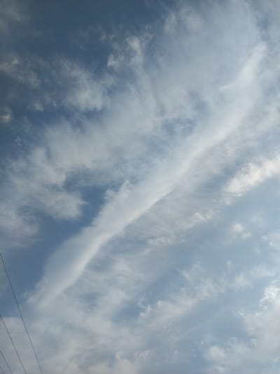 The image shows a sky with scattered clouds. Three dark, thin lines, likely power lines, run diagonally from the lower left corner upwards. The sky is blue with varying shades of white clouds, some wispy and some more dense. A long, thin cloud formation stretches diagonally across the upper half of the frame. The scene appears to be outdoors, with no visible people or specific landmarks. The environmental conditions suggest daytime, possibly with some atmospheric haze or high cloud cover.